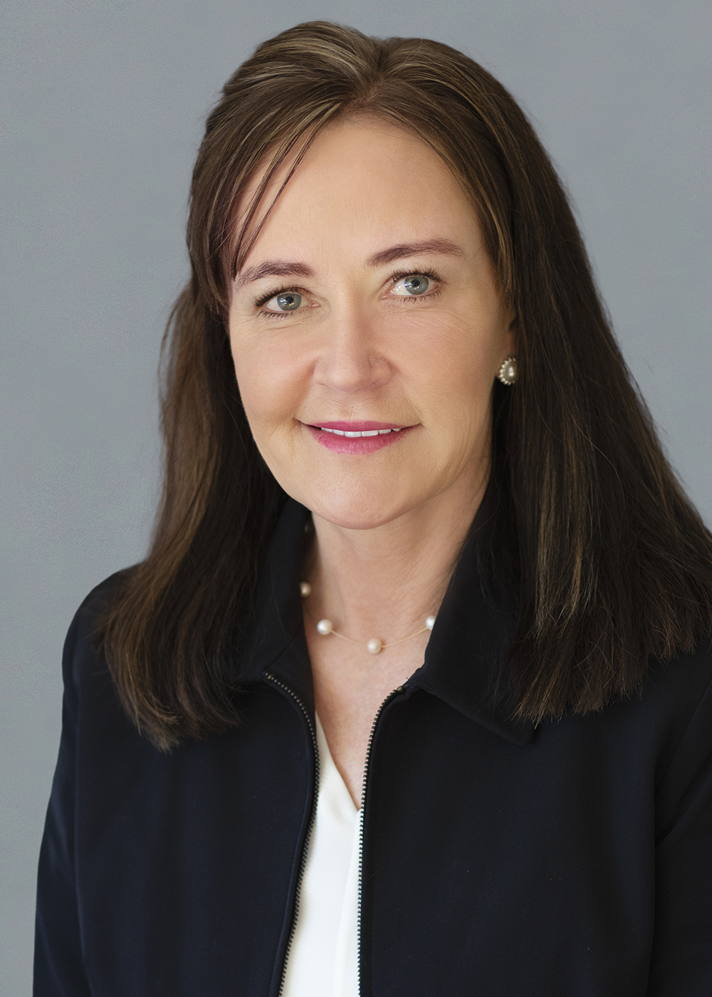 Professional woman with a confident smile wearing a dark jacket and a pearl necklace against a grey background.