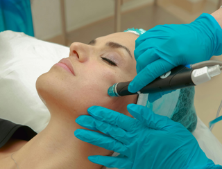 A woman lies with eyes closed while a professional wearing blue gloves performs a facial treatment using a handheld device; she wears a disposable cap in a clinical setting.