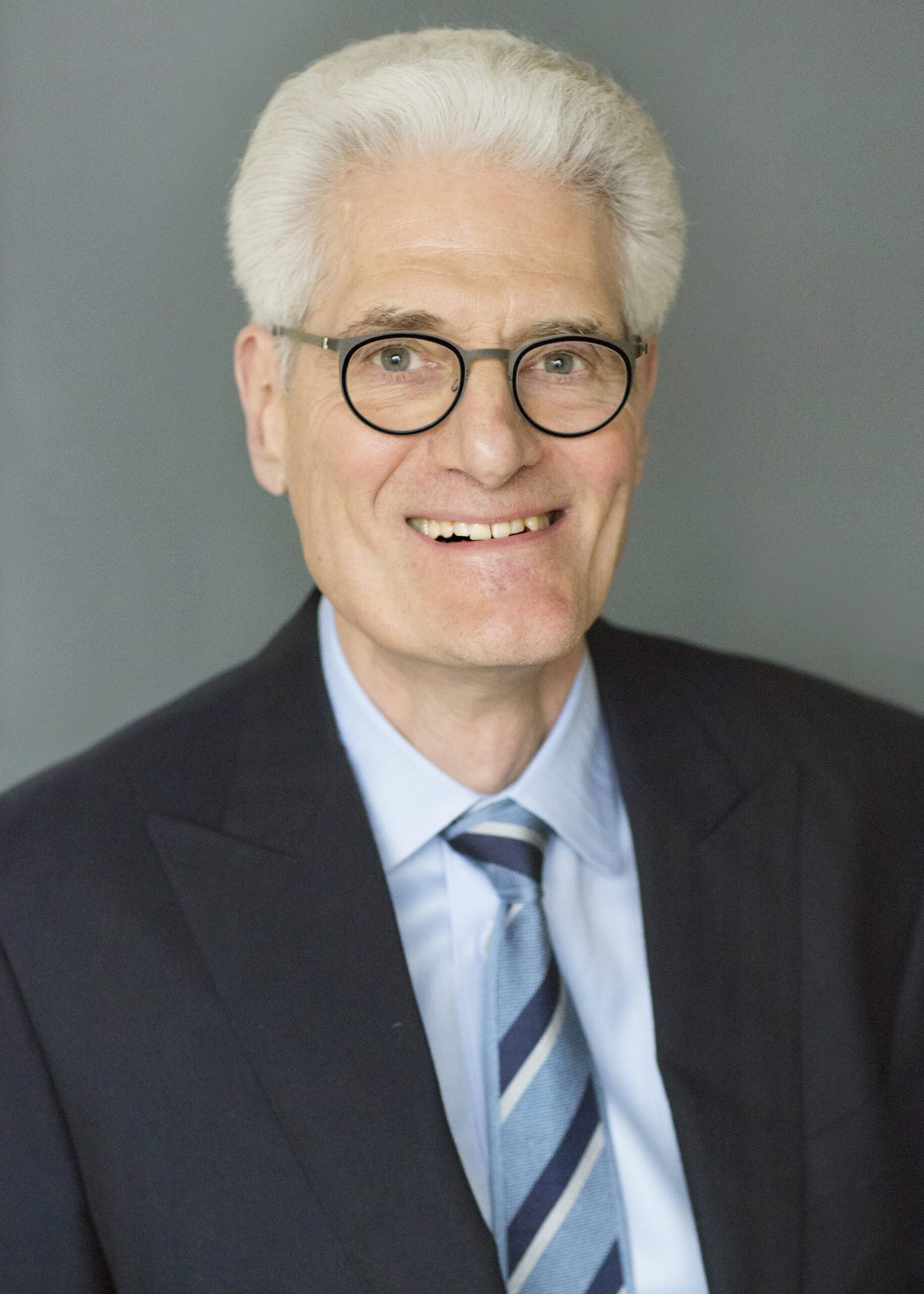 A professional portrait of a smiling senior man with silver hair, wearing glasses, a blue suit, a light blue shirt, and a striped tie.