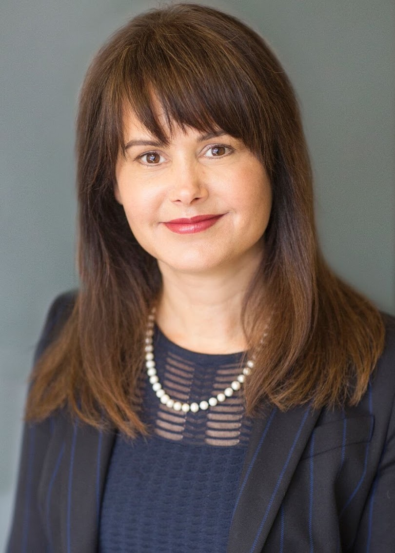 A professional portrait of a woman with shoulder-length brown hair, smiling gently at the camera, wearing a dark blazer and a necklace, set against a neutral background.