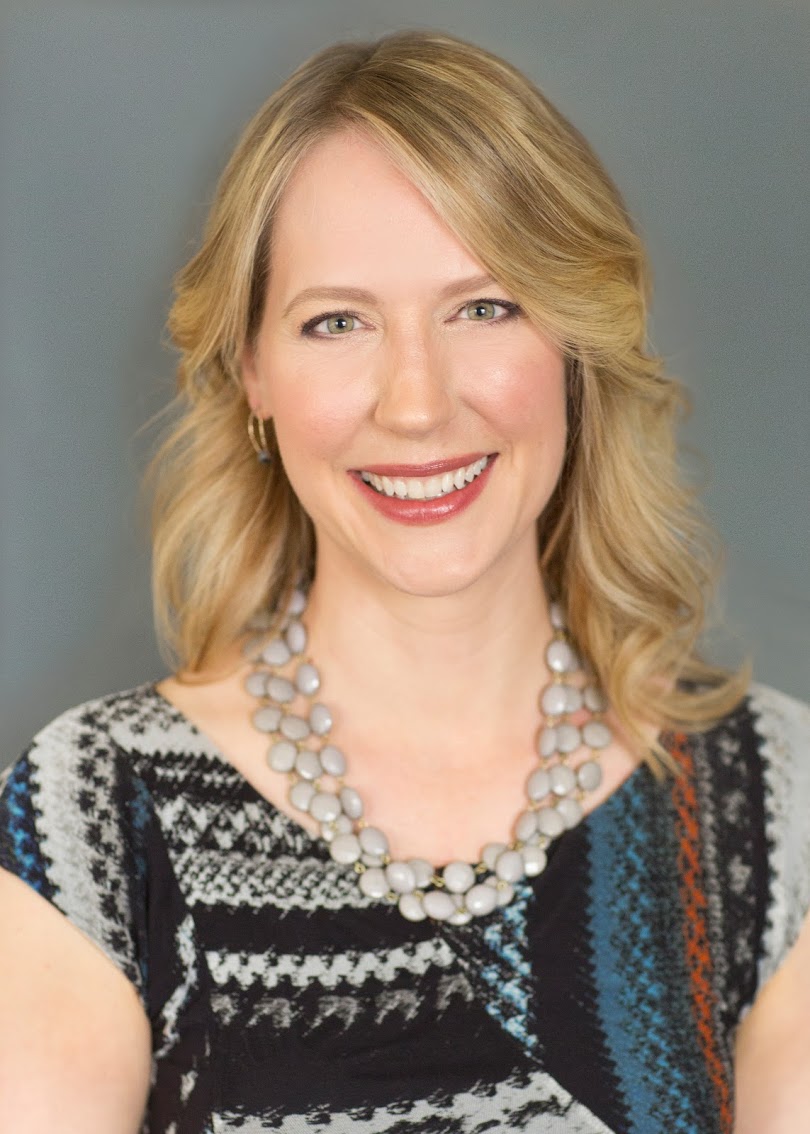 Professional portrait of a smiling woman with blonde hair, wearing a patterned dress and a pearl necklace.