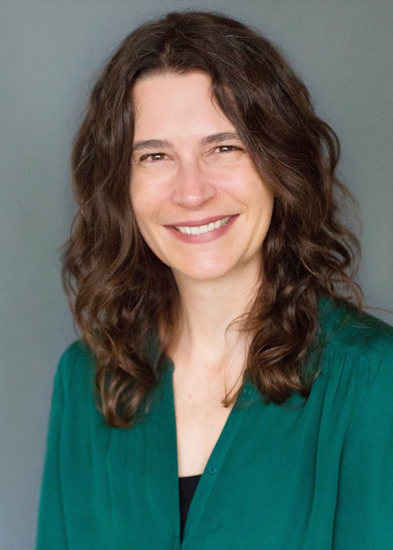 A smiling woman with long wavy hair wearing a green blouse against a neutral gray background.