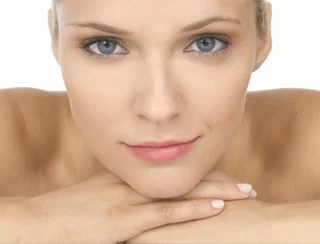 A close-up of a woman with clear blue eyes and a subtle smile, resting her chin on her hands.