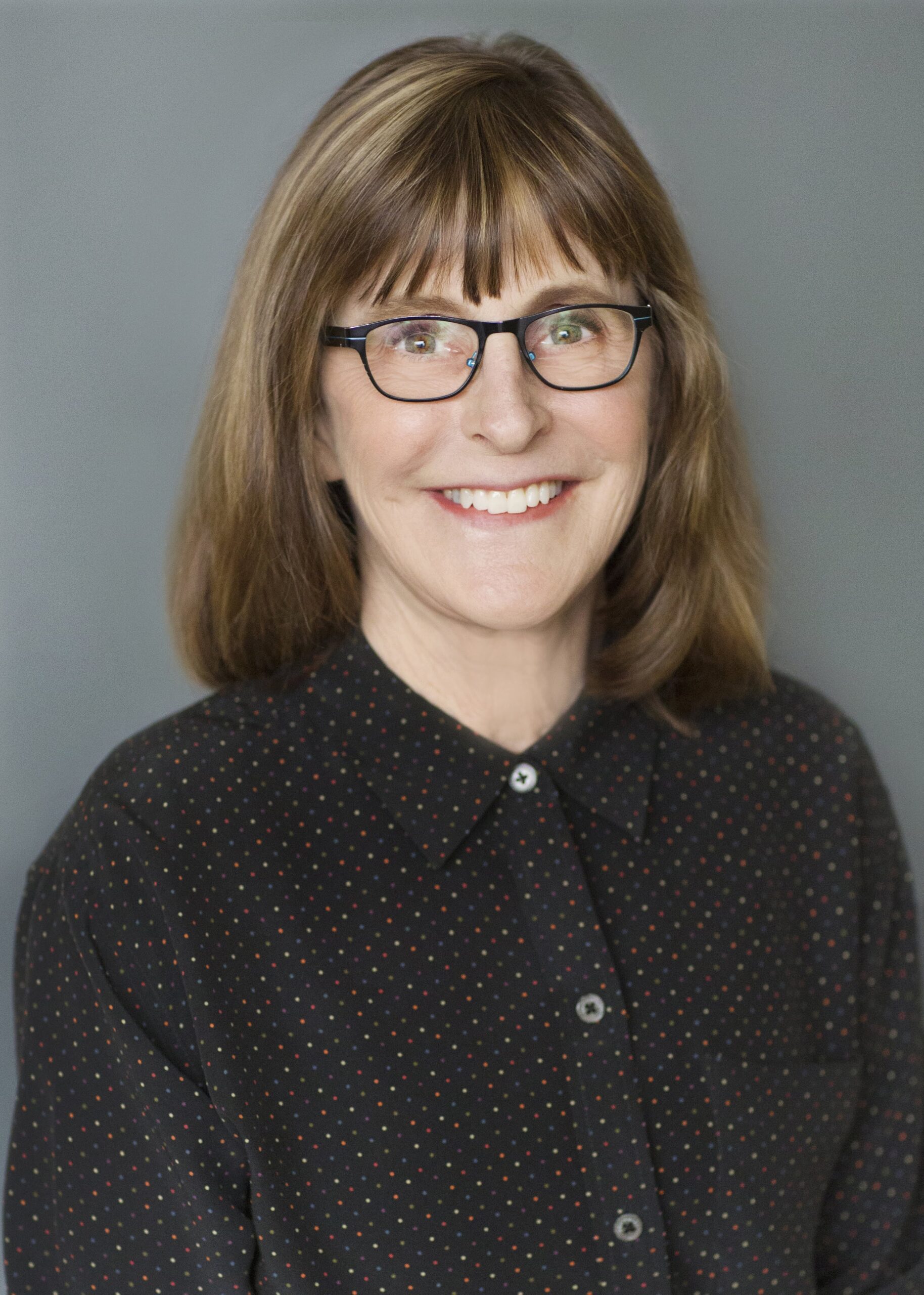 A smiling woman with glasses wearing a dotted blouse against a neutral background.