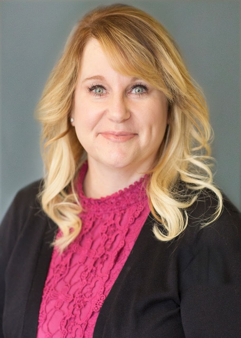 Professional headshot of a woman with blonde hair and blue eyes, wearing a pink blouse with a lace collar, exuding confidence and approachability.