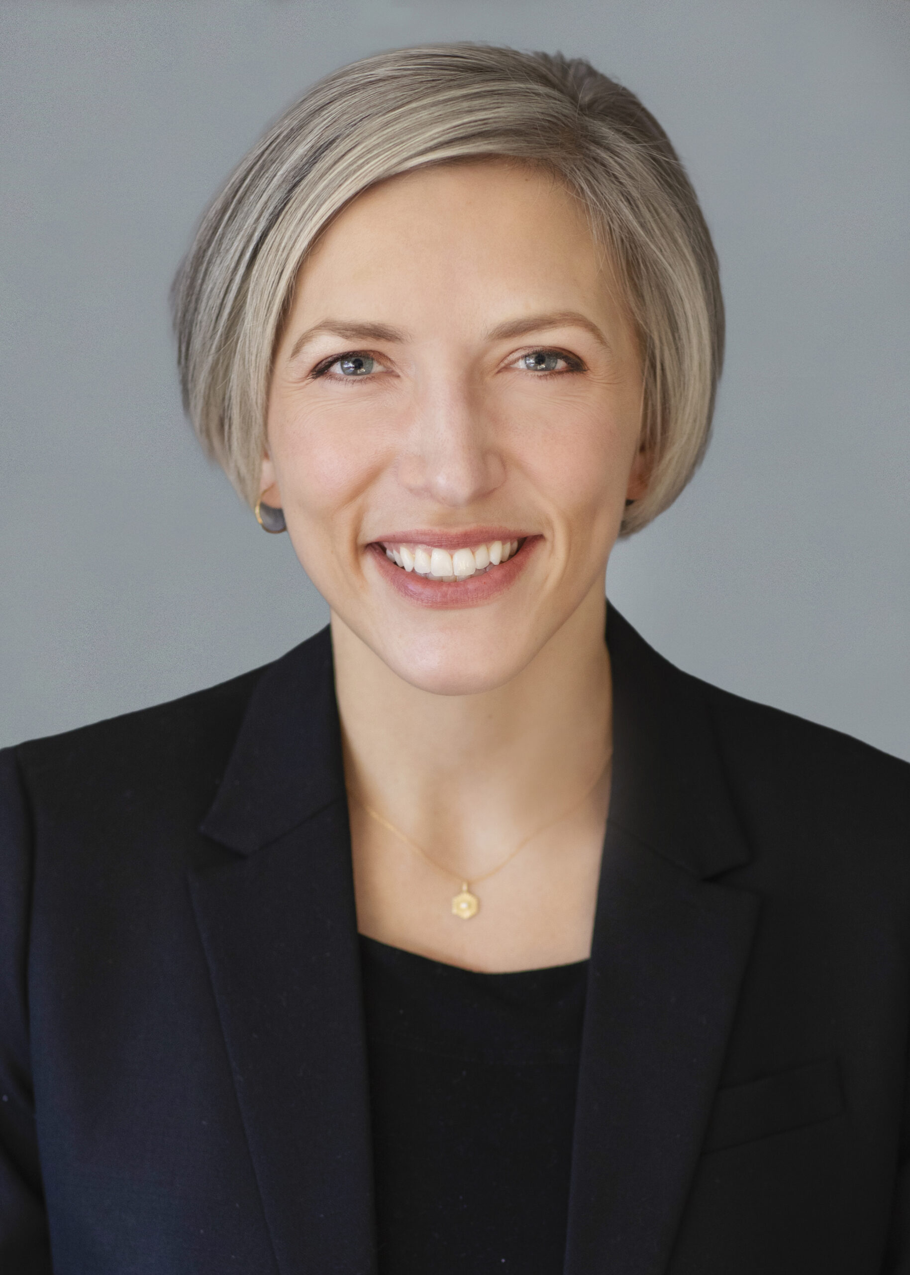 A professional headshot of a smiling woman with short silver hair, wearing a dark blazer and a small pendant necklace, set against a light blue background.