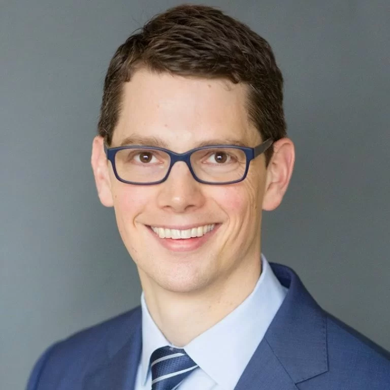 Smiling young professional wearing blue-framed glasses and a sharp blue suit with a striped tie, projecting confidence and approachability.