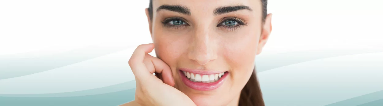 Portrait of a smiling woman with dark hair and clear skin, resting her face on her hand against a light background.