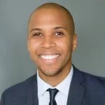 A professional portrait of a smiling person with a clean haircut, wearing a suit and tie.