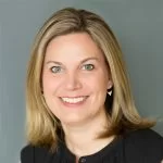 A professional portrait of a smiling woman with shoulder-length blond hair, wearing a black top and small earrings, set against a grey background.