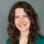 A smiling woman with shoulder-length curly hair, wearing a green top and displaying a friendly demeanor against a neutral background.