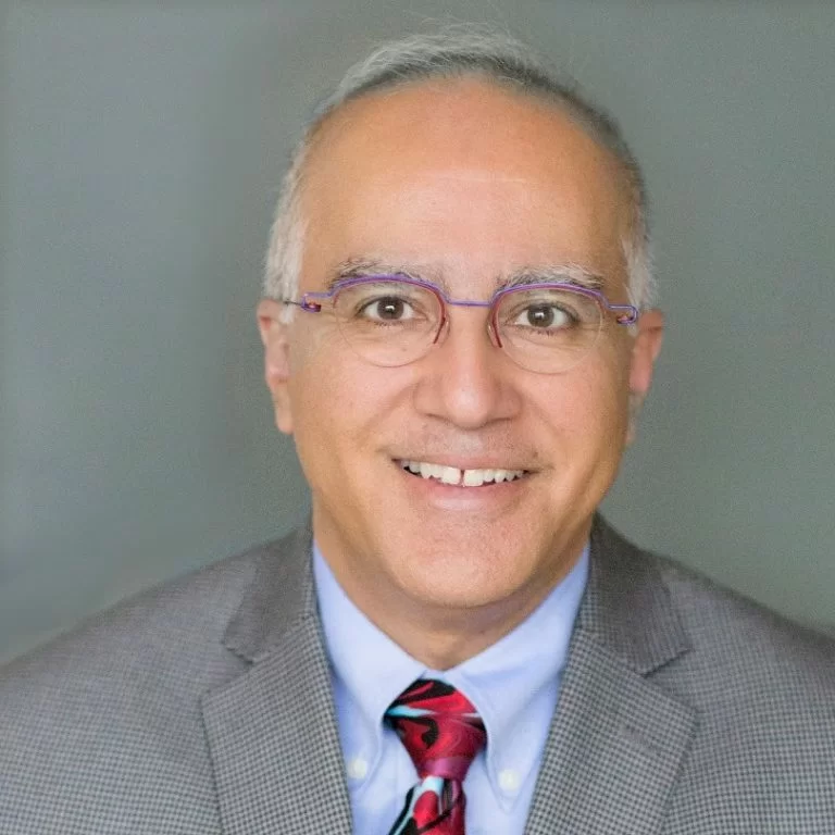 A professional portrait of a smiling man with glasses, wearing a patterned tie and a gray suit jacket.