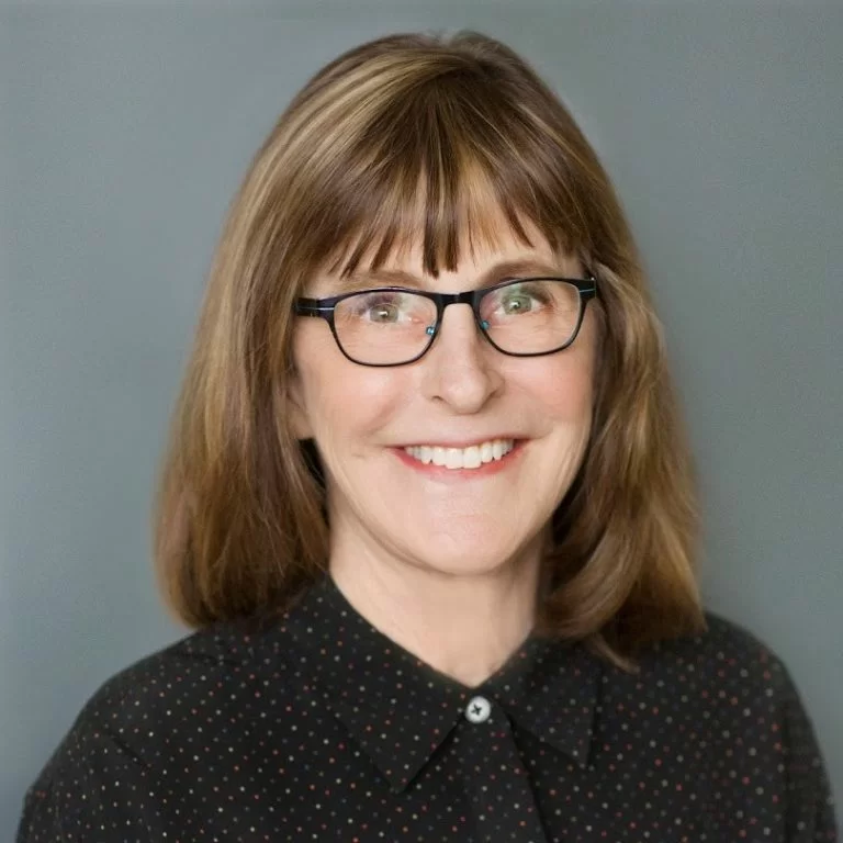 A smiling woman with glasses, medium-length hair, and a polka-dotted blouse against a neutral background.