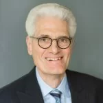 Mature professional man with a warm smile, wearing glasses, a suit, and a light blue tie.