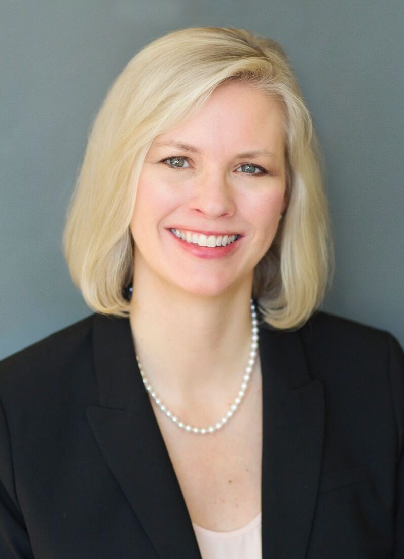 A professional woman with a confident smile, wearing a black blazer and a pearl necklace, posing for a formal portrait against a grey background.