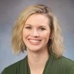 A smiling woman with short blonde hair wearing a green top against a grey background.