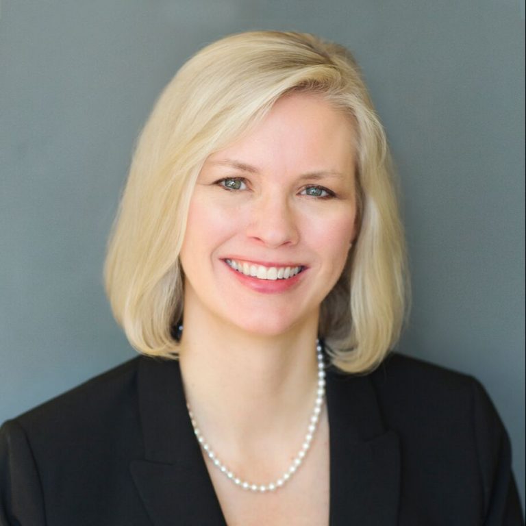 A professional portrait of a smiling woman with blond hair, wearing a black blazer and a string of pearls.