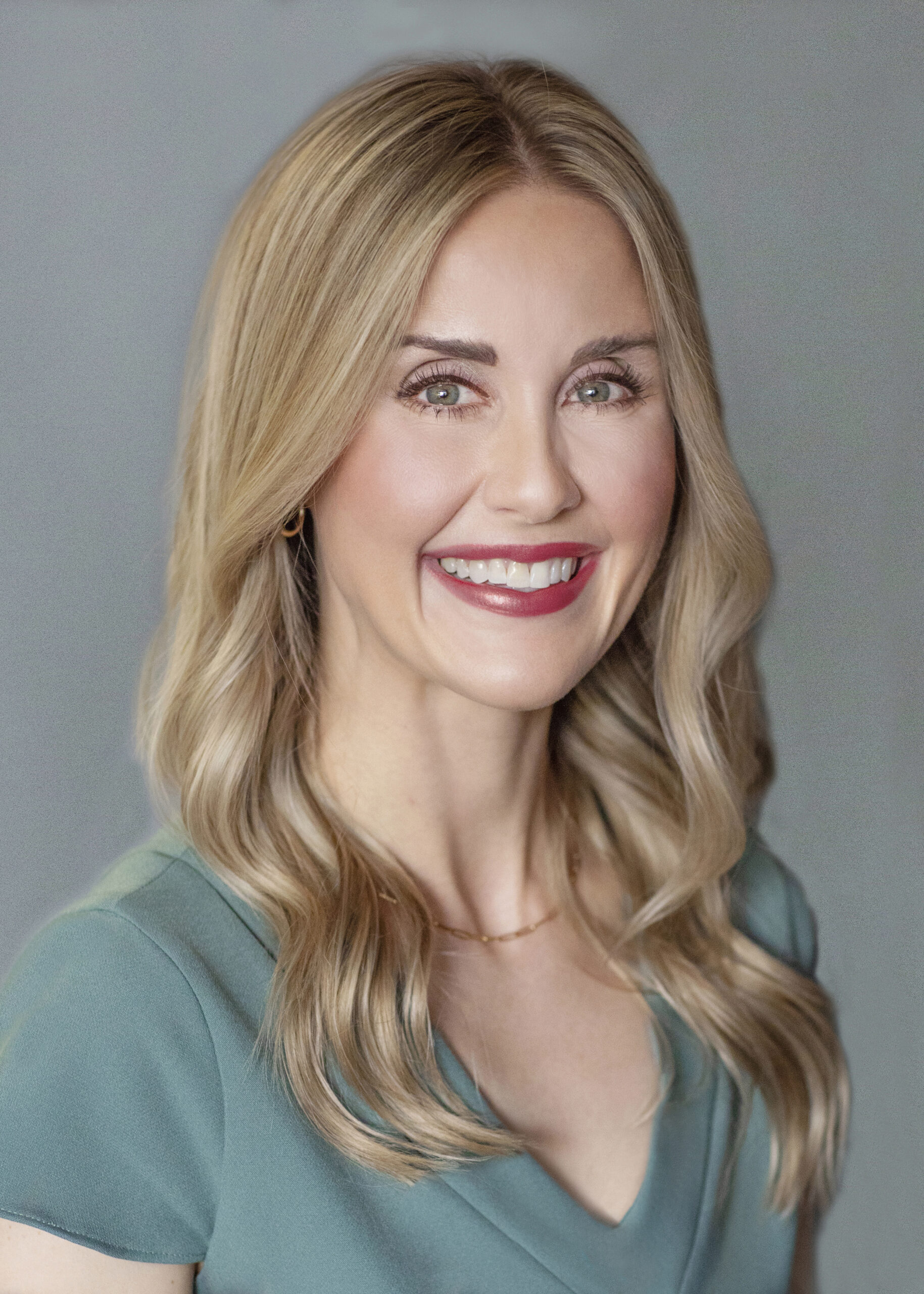 A professional headshot of a smiling woman with blonde hair wearing a teal top.