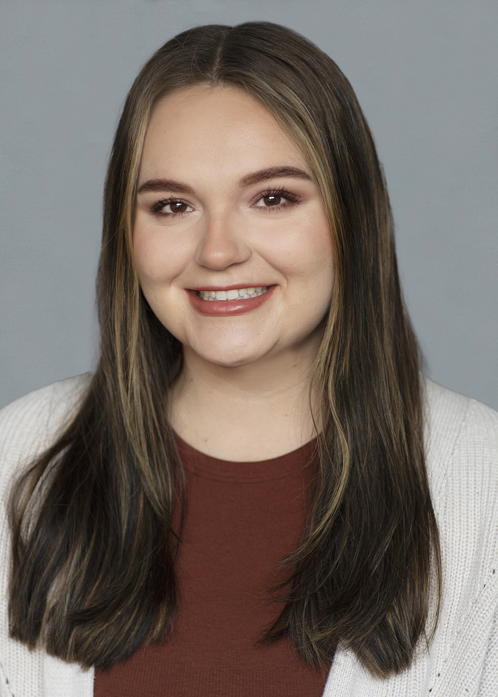 A smiling young woman with shoulder-length brown hair, wearing a burgundy top and a white cardigan, against a gray background.