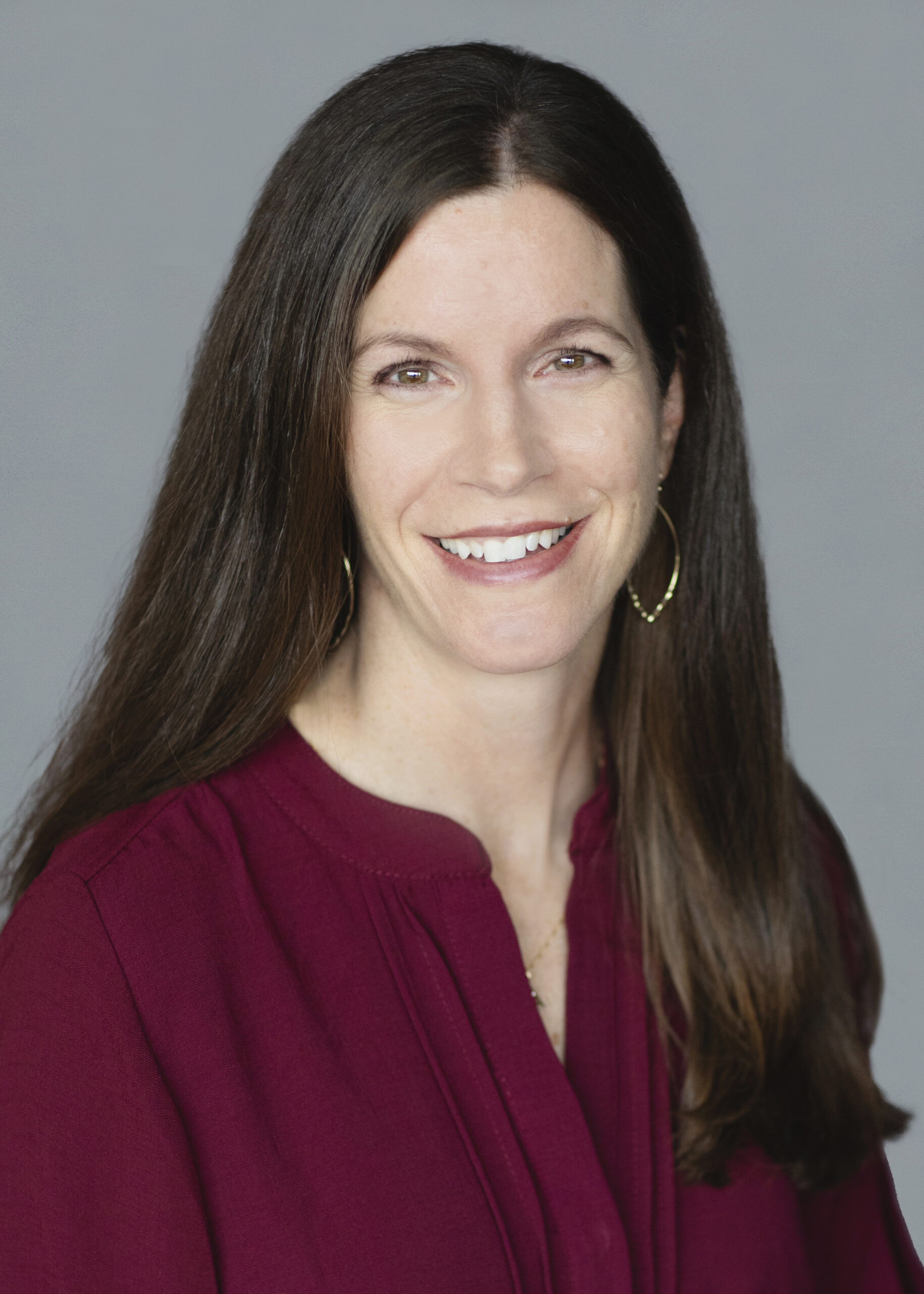 Professional woman with a friendly smile, wearing a burgundy blouse and hoop earrings, against a gray backdrop.