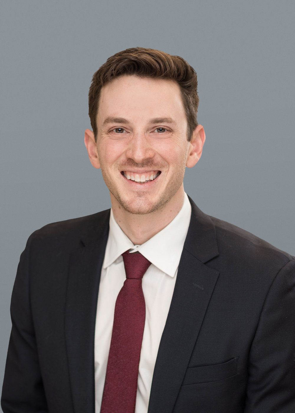 A man in a dark suit, white shirt, and maroon tie smiles at the camera against a plain gray background.