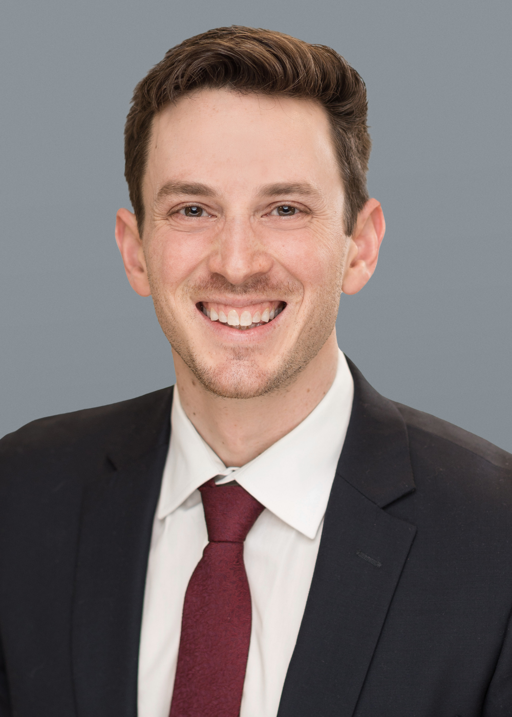 A man in a dark suit, white shirt, and burgundy tie is smiling at the camera against a plain gray background.