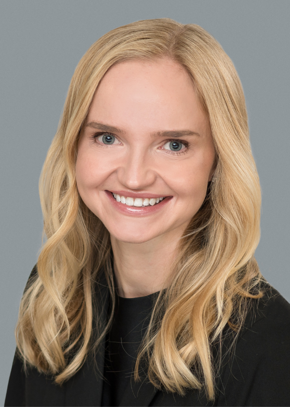 A woman with long, wavy blonde hair smiles at the camera. She is wearing a black top and jacket, and is posed in front of a plain gray background.