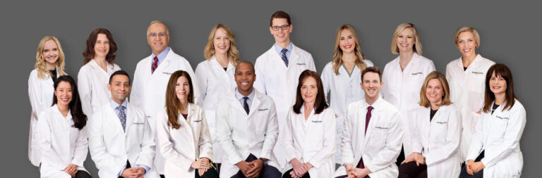 A group of seventeen medical professionals, wearing white lab coats and smiling, pose in two rows against a plain gray background.