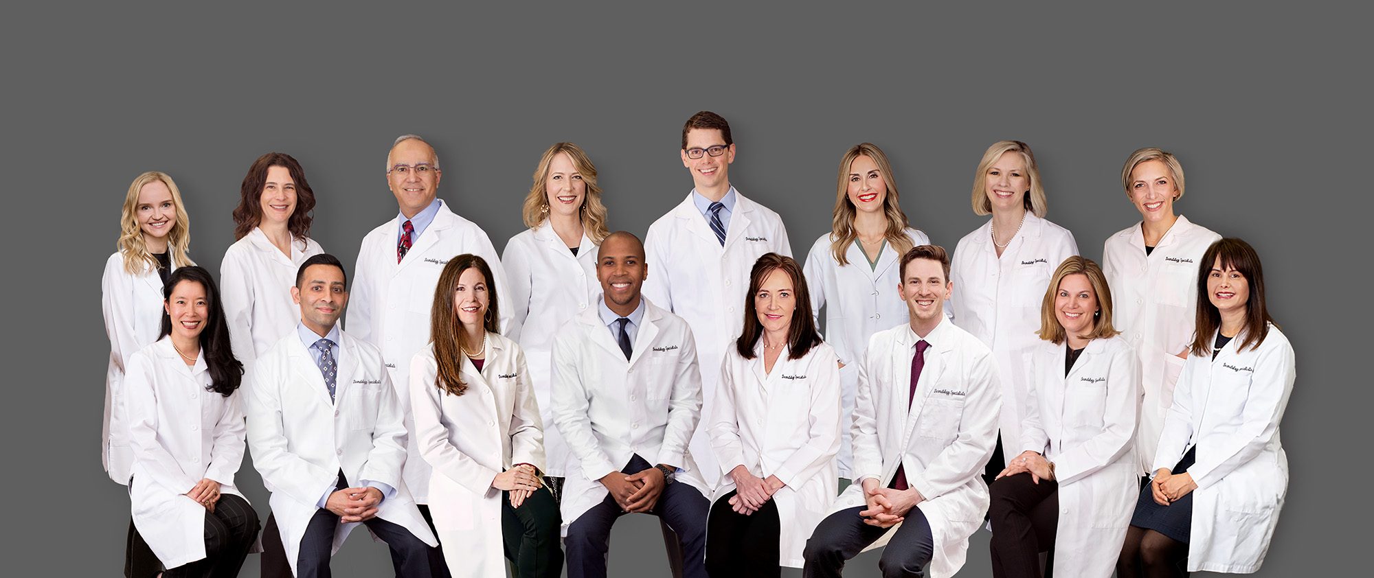 A group of fifteen medical professionals, wearing white lab coats, pose together and smile in front of a plain gray background. Eight are seated in the front row and seven are standing behind them.