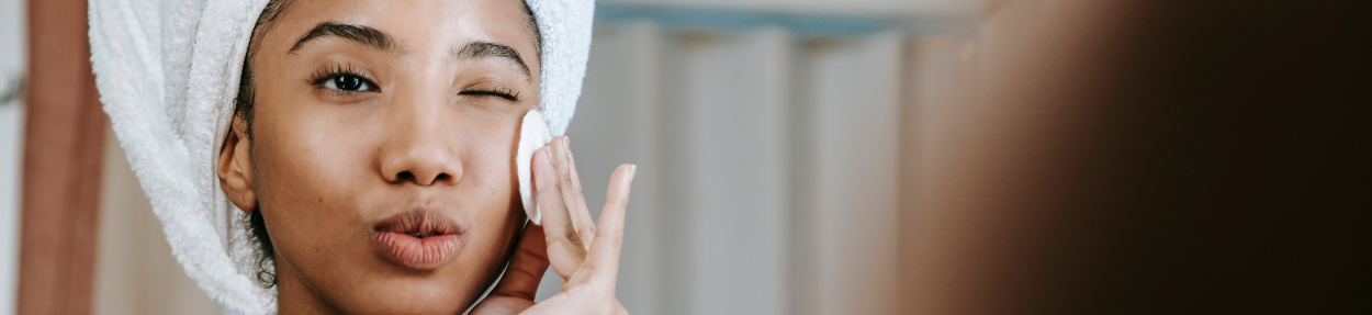 Untitled design - 2025-10-15T201105.538 A woman with a white towel wrapped around her head applies skincare cream to her face, winking with one eye and pursing her lips in front of a bathroom mirror.