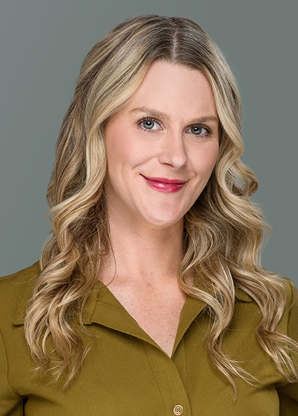A woman with long blonde wavy hair and blue eyes smiles softly. She is wearing an olive green top and stands in front of a plain gray background.