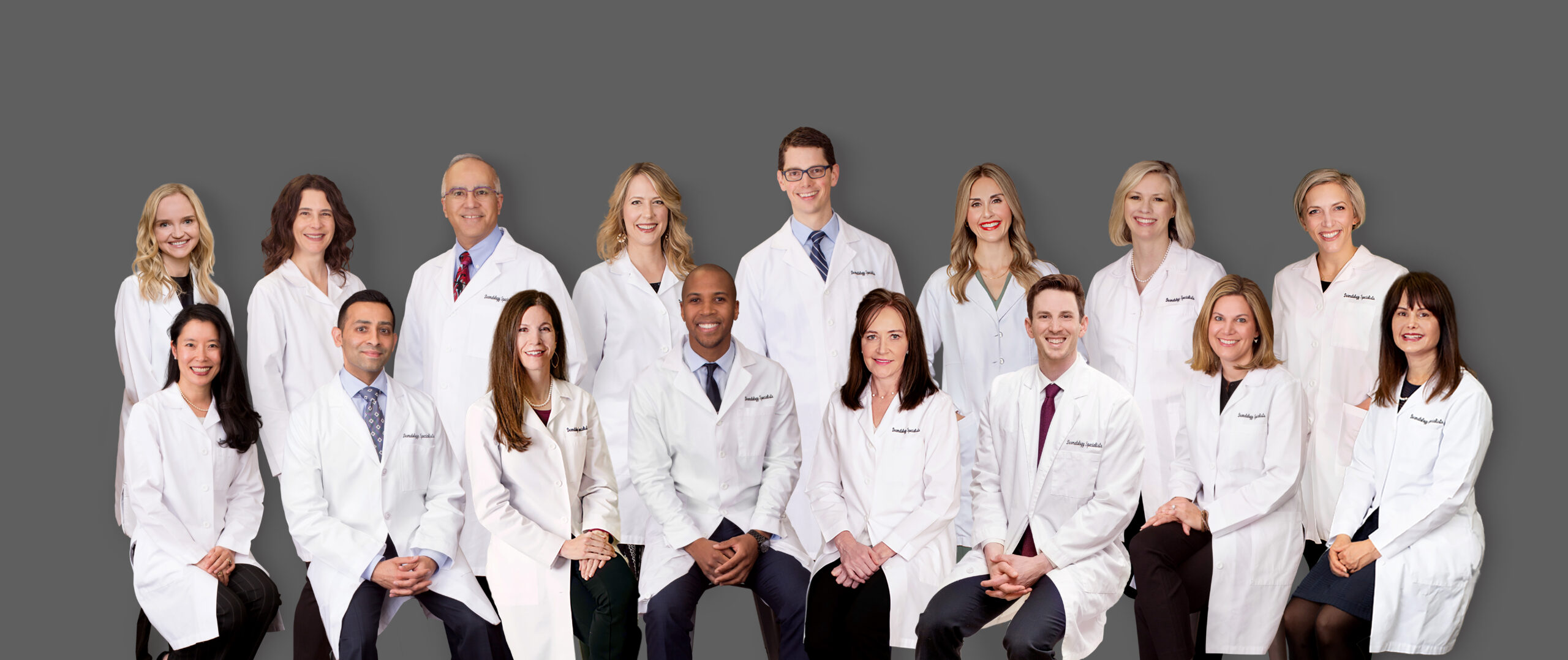A group of fifteen medical professionals, dressed in white lab coats, sit and stand together against a plain gray background, smiling and posing for a group photo.